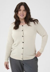 Bež kardigan s okroglim izrezom, dolgimi rokavi in šestimi črnimi gumbi spredaj. Mehak material, prilegajoča zasnova, v kombinaciji s sivo hlačami.