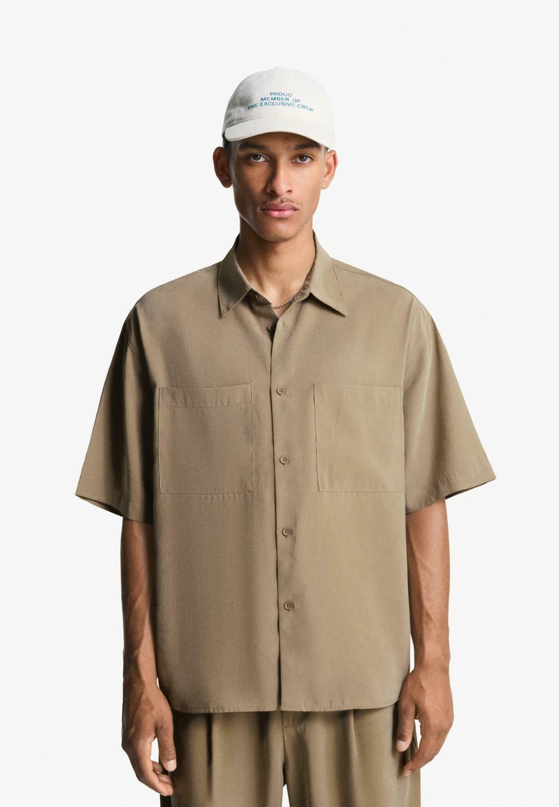 Korte mouwen, beige knoopshirt met twee voorkruiszakken, gemaakt van gladde stof, gecombineerd met een lichtgekleurde, geborduurde pet.