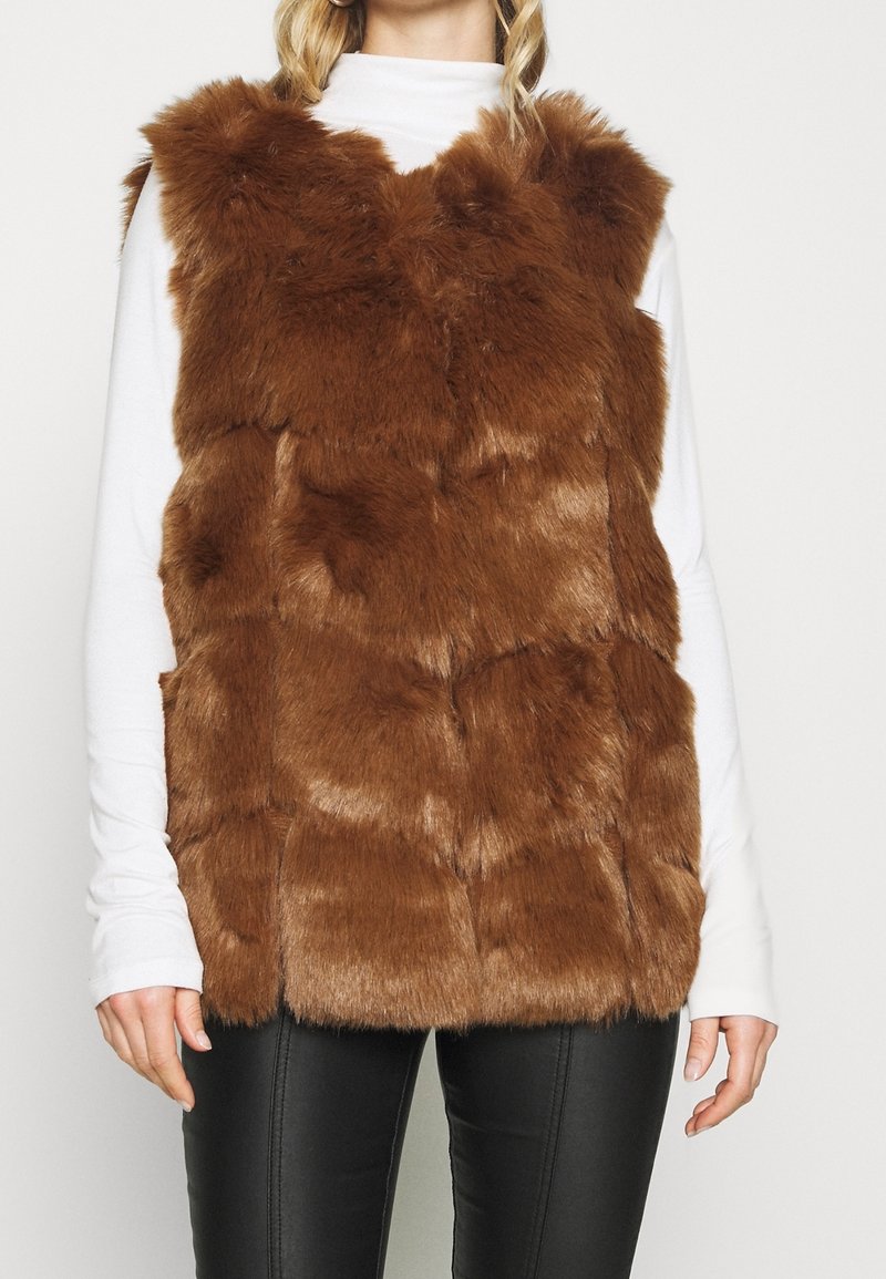 Gilet in pelliccia sintetica marrone con design a trama trapuntata. Senza maniche, con scollo alto, indossato sopra una maglietta bianca a maniche lunghe, abbinato a pantaloni neri.