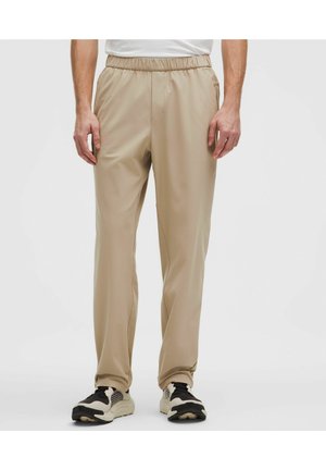 Uomo che indossa pantaloni beige con vita elasticizzata e gamba dritta e sneakers bianche e nere, in piedi davanti a uno sfondo chiaro e uniforme.