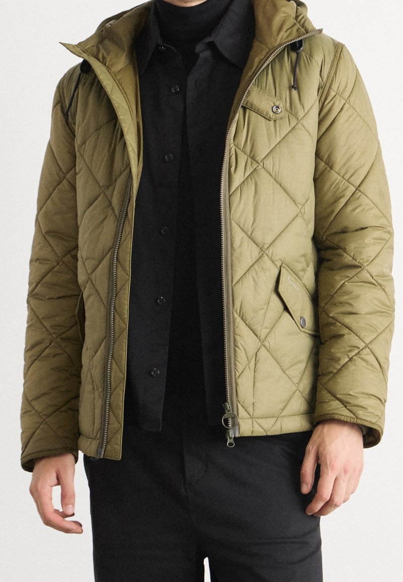 Gestepptes grünes Jacke mit durchgehendem Reißverschluss, Kapuze und Seitentaschen. Hat ein diagonales Muster und schwarze Knopfakzente. Getragen über einem schwarzen Oberteil.