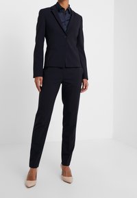 Costume bleu marine sur mesure avec une veste cintrée et un pantalon droit. Le tissu semble lisse, associé à une chemise sombre et des talons beiges.