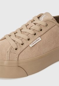 Beige suede sneaker featuring round toe, lace-up front, tan rubber sole, and a small Calvin Klein logo tag on the side.