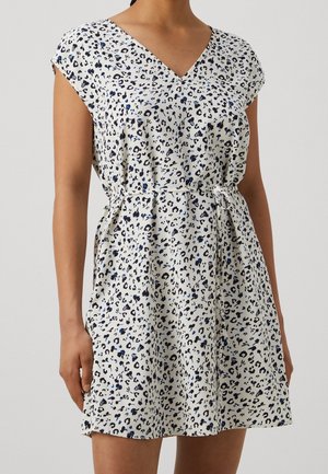 Vrouw draagt een wit jurkje met korte mouwen, V-hals, zwart- en blauw luipaardprint en een ceintuur in de taille.