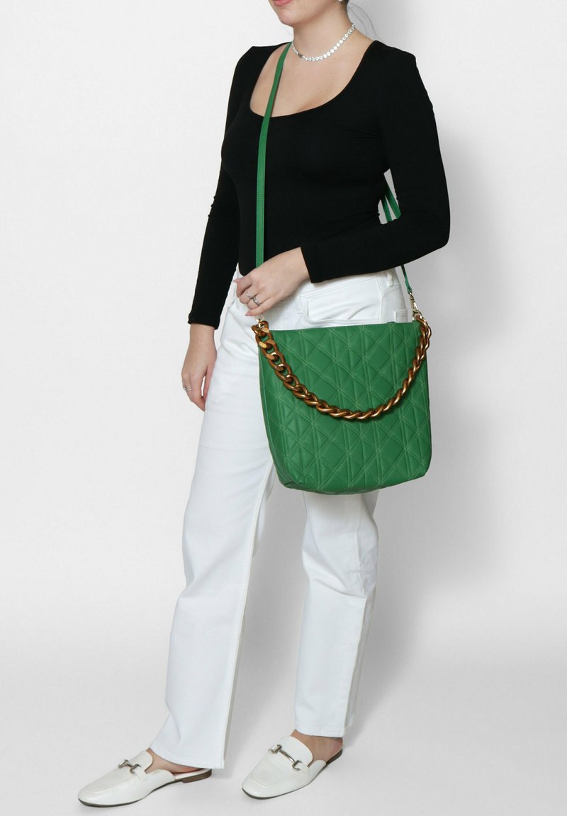 Borsa a tracolla verde trapuntata con accenti in oro, caratterizzata da una forma strutturata e una tracolla regolabile. Indossata da una persona con una maglietta nera e pantaloni bianchi.