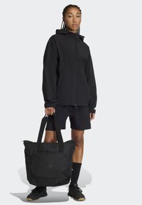 Czarna wodoodporna kurtka na zamek, zestawiona z czarnymi szortami i pasującą torbą na ramię. Torba ma dwa paski na ramię oraz przednią kieszeń.