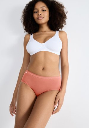 Vrouw met krullend haar die een witte beha en koraalkleurige hipsteronderbroek draagt, staand tegen een effen lichte achtergrond.