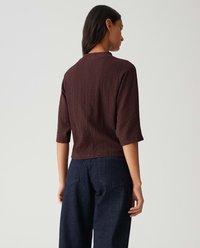 Eine Frau von hinten zu sehen, die ein strukturiertes, dunkelbraunes Oberteil mit Ellenbogenlängenärmeln und dunkelblaue, hochtaillierte, weite Jeans trägt.