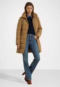 Barna puffermellény magas gallérral, cipzáras zsebekkel és térd fölé érő kivitelben. Sötétkék pulóver és kék farmer nadrág felett viselve. Barna magassarkú csizmák.