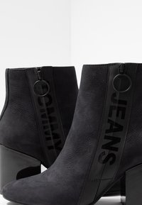 Botas negras hasta el tobillo con textura de gamuza, acentos brillantes y un tacón bloque pronunciado. Presenta un audaz logo de "TOMMY JEANS" en los laterales.