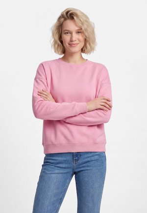 Jeune femme aux cheveux blonds courts portant un sweat-shirt rose et un jean bleu, debout les bras croisés, souriant légèrement.