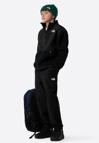 Čierna flísová bunda so zipsom, čierne nohavice, zelená pletená čiapka a batoh. Na bunde a nohaviciach je logo. Textúrované detaily látky.