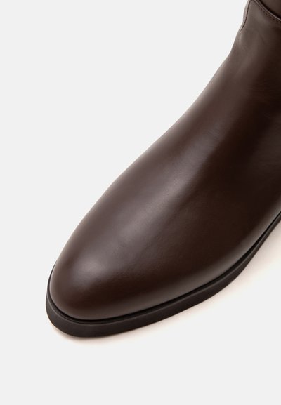 Bout de chaussure en cuir marron foncé avec une finition lisse et une semelle noire sur un fond clair uni.