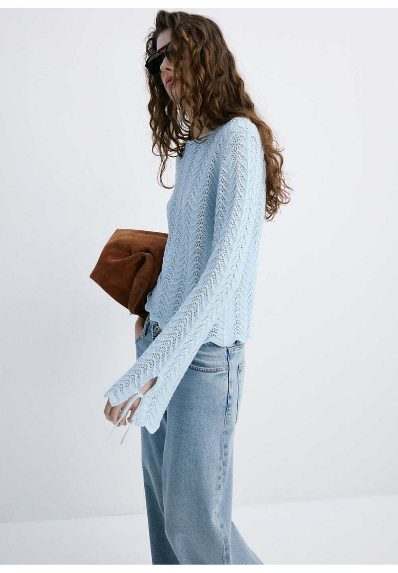 Femme aux longs cheveux bouclés portant des lunettes de soleil noires, un pull en tricot bleu clair, un jean bleu ample, tenant une grande pochette marron.