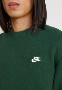 Πράσινο πλεκτό πουλόβερ με ριχτό λαιμόκοψο και μικρό λευκό λογότυπο Nike στο στήθος. Μαλακή υφή και άνετη εφαρμογή.