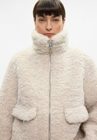 Beež faux shearling jakk, millel on kõrge kaelus, lukuga sulgemine ja kaks eesmist taskut. Pehme, tekstuuriga kangas loob hubase väljanägemise.