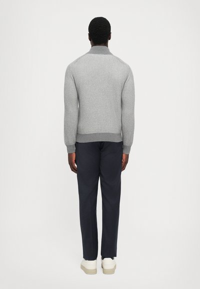 Grau gerippter Pullover mit hohem Kragen, kombiniert mit marineblauen Hosen und weißen Sneakers, von hinten sichtbar. Texturdetail des Stoffes ist sichtbar.