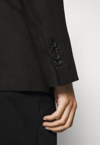 Manche d'un blazer noir cintré avec quatre boutons noirs, tissu texturé et coupe élégante. Main reposant sur le côté.