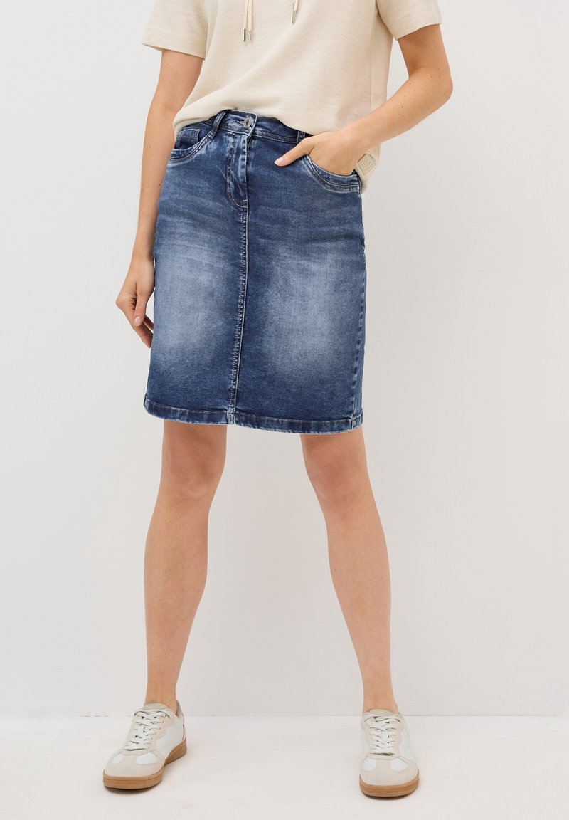 Femme portant une jupe en jean bleu délavé longueur genou, un haut à manches courtes écru et des baskets blanches, debout avec une main dans la poche.