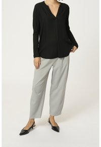 Kvinde iført sort V-hals bluse med lange ærmer, lysegrå skræddersyede bukser og sorte slingback-sko med spids tå, stående foran en ensfarvet baggrund.