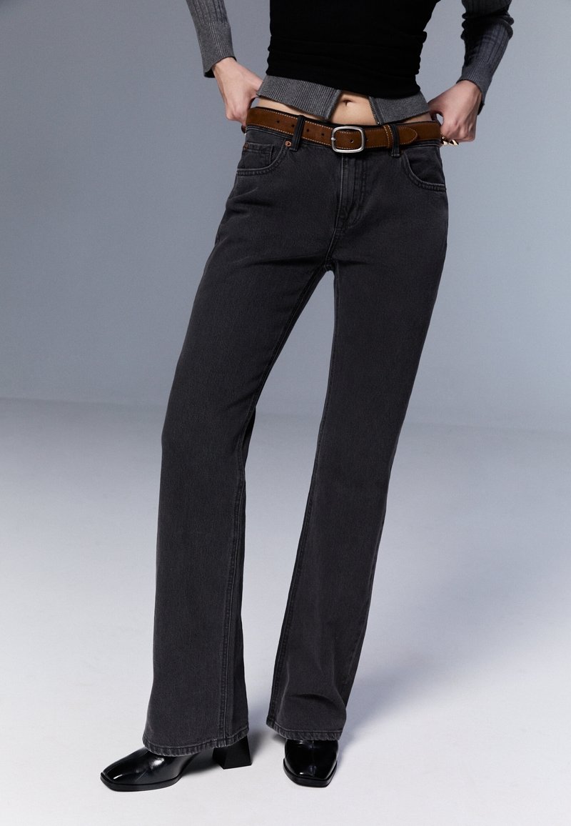 Personne portant un jean noir taille haute, une ceinture marron, des chaussures noires à bouts carrés et un haut gris à manches longues, remontant le jean à la taille.