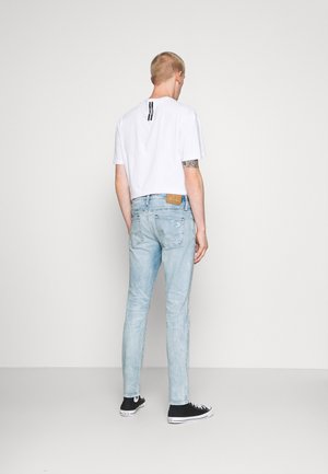 Junger Mann mit weißem T-Shirt, hellblauen Jeans und schwarzen Turnschuhen, der mit dem Rücken zur Kamera vor einem schlichten weißen Hintergrund steht.