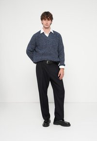 Tumši zila adīta džemperis ar V-veida izgriezumu, valkāts virs baltas kreklas ar apkakli; kopā ar tumšām, pielāgotām biksēm un melniem apaviem.