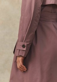 Detailný záber predlaktia osoby v jemnej marhuľovej trench coat, so zapínaním na gombíky a viditeľnými stehmi.