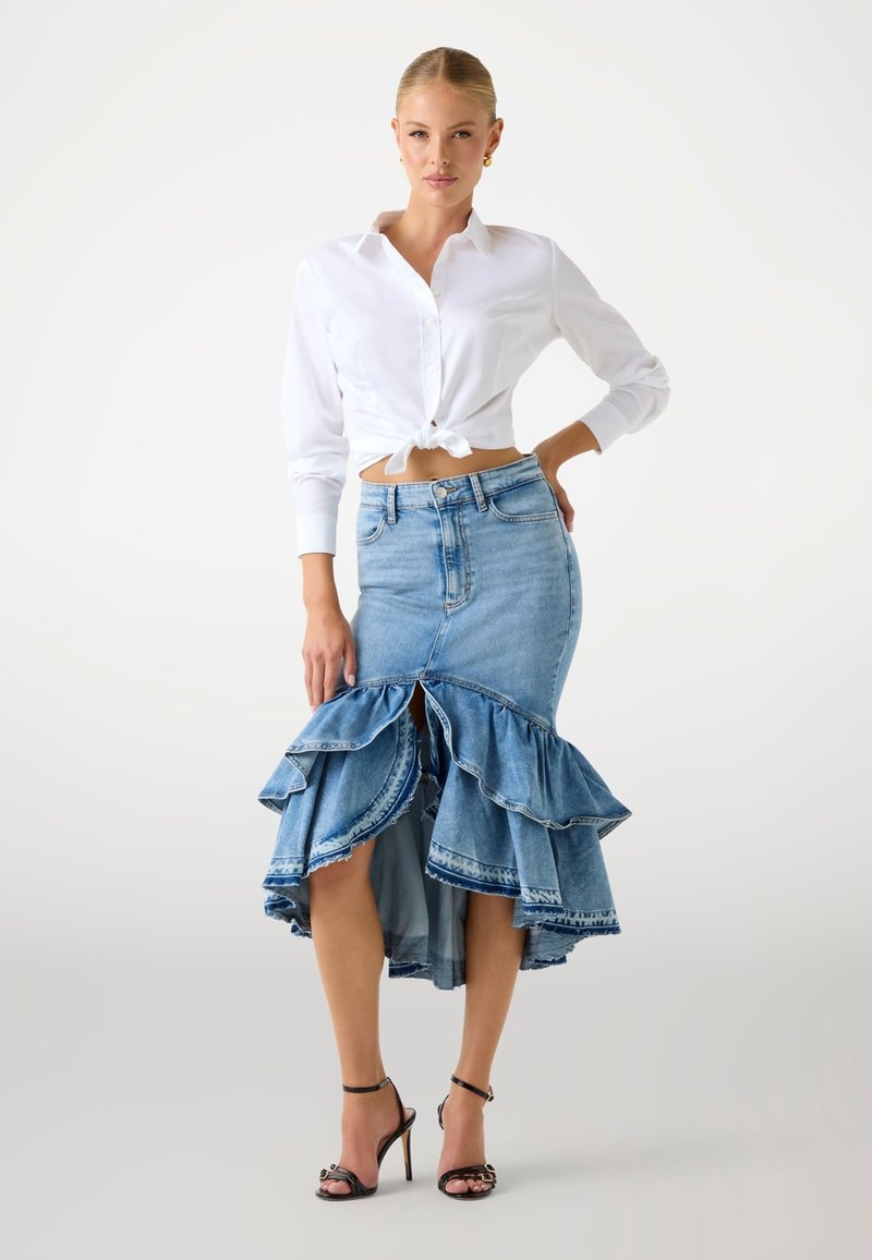 Vrouw in een geknoopte witte blouse, asymmetrische denim rok met ruches en zwarte hoge hakken, staande tegen een effen achtergrond.