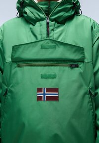 Vihreä pullover-takki hupulla, jossa on etutasku, vaakasuora vetoketju ja Islannin lipun merkki. Rakenne on teksturoitu ja vedenpitävä.