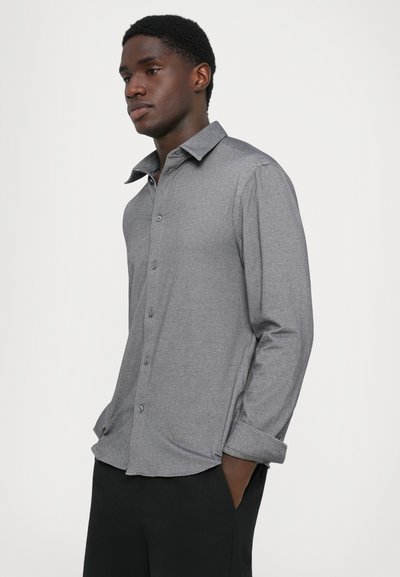 Giovane con le mani nelle tasche, che indossa una camicia a maniche lunghe grigia con bottoni e pantaloni neri, che guarda di lato su uno sfondo bianco.