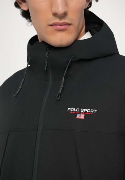 Jachetă ușoară de culoare neagră cu glugă, având un fermoar frontal și șnur de ajustare. Logo-ul brodat "POLO SPORT" în roșu și alb pe piept.