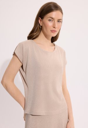 Kobieta o jasnobrązowych włosach ubrana w krótkim rękawem, błyszczącą beżową bluzkę i dopasowaną spódnicę, stoi z jedną ręką na biodrze.