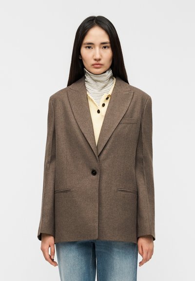 Blazer marron sur mesure avec un bouton unique, deux poches avant et un col à revers en encoche, porté sur un haut de couleur crème et un col roulé rayé.