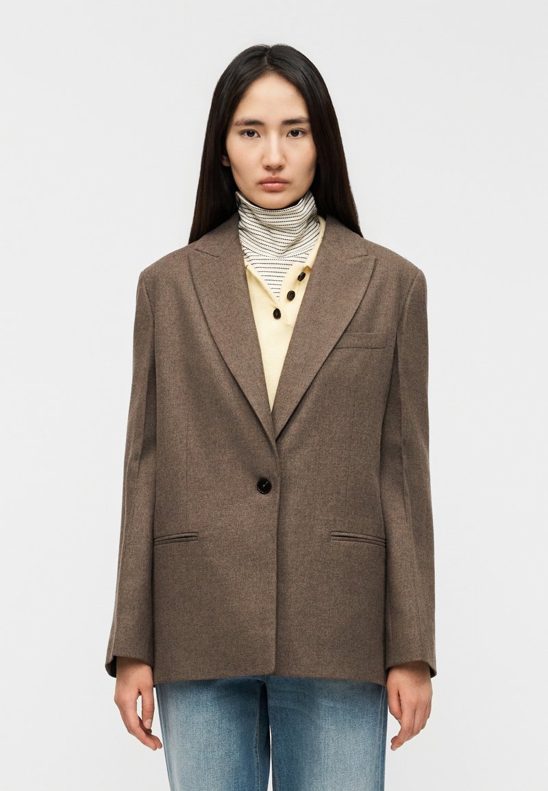 Καφέ σακάκι ραμμένο κατά παραγγελία με ένα κουμπ ί, δύο μπροστινές τσέπες και γιακά με χαραγή, φορεμένο πάνω από μια μπλούζα εκρού χρώματος και ριγωτό ζιβάγκο.