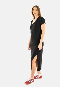 Robe noire à manches courtes, décolleté en V et fente sur le côté. Fabriquée en tissu doux, avec un branding minimal sur la poitrine. Associée à des baskets rouges et blanches.