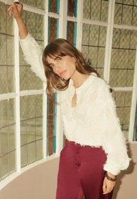 Bela teksturirana bluza z resicami, z gumbi za zapenjanje in dolgimi rokavi. Kombinirana z visokim pasom bordo hlač. Naravna svetloba.