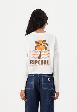 Crop top blanc à manches longues avec un graphique coloré d'un palmier, d'un soleil et de vagues, portant la marque "RIPCURL" en lettres brunes en gras. Pantalons en denim bleu.