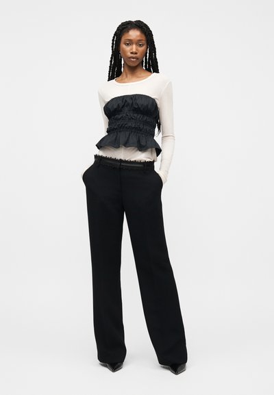 Γυναίκα με μακριά κοτσίδα που φοράει λευκή μπλούζα με μακριά μανίκια, από πάνω ένα μαύρο μπούστο με ρυτίδες, μαύρο παντελόνι με φαρδιά μπατζάκια και μαύρα μυτερά παπούτσια.