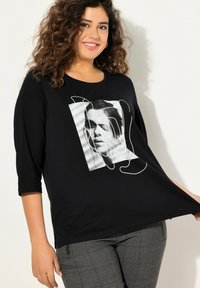 Maglietta grafica nera con scollatura rotonda, maniche a tre quarti, presenta un disegno a linee bianche su uno sfondo fotografico. Pantaloni a quadri grigi texture.