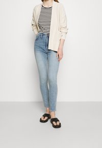 Světle béžový kardigan přes černý a bílý pruhovaný top, spárovaný s úzkými světle modrými džíny a černými nazouvacími sandály.