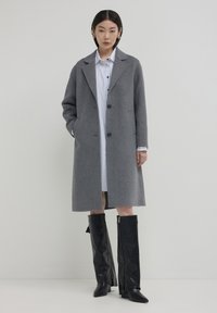 Cappotto grigio di lana con revers, due tasche anteriori e bottoni neri, abbinato a una camicia a righe bianche e stivali neri al ginocchio.