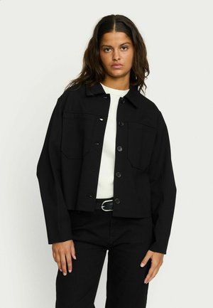 Schwarze, cropped Jacke aus Stoff mit einer vorderen Knopfleiste und zwei Brusttaschen. Getragen über einem weißen Oberteil und kombiniert mit schwarzen Hosen.