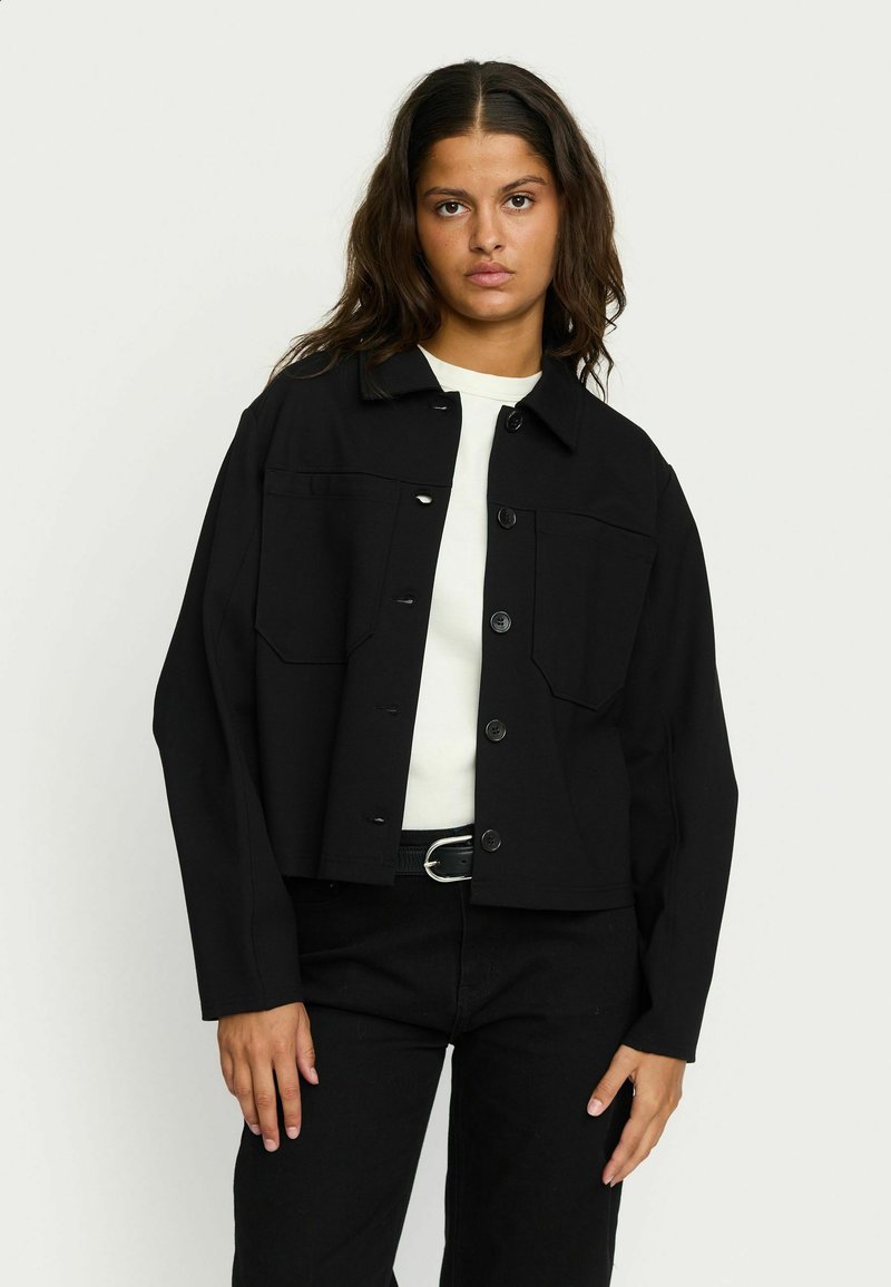 Zwart cropped jack van stof met een sluiting met een knop aan de voorkant en twee borstzakken. Draag het over een witte top en combineer het met zwarte broek.