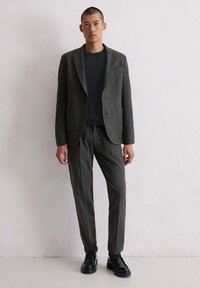 Man in een donkergrijze blazer en bijpassende broek met zwarte schoenen, staand tegen een lichtgrijze gestructureerde muur.