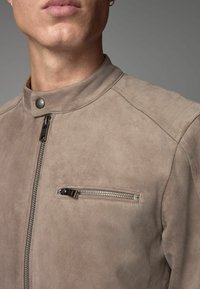 Chaqueta de ante beige con un alto cuello, que presenta un bolsillo en el pecho con cremallera y detalles de costura sutiles en los hombros. Textura suave.