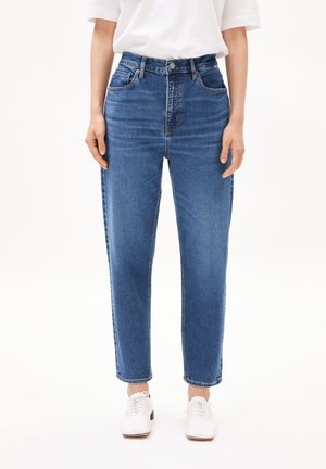 Person trägt mittelhoch sitzende, gerade geschnittene blaue Jeans in Kombination mit weißen Schnürschuhen und einem in die Hose gesteckten weißen kurzärmligen Hemd.