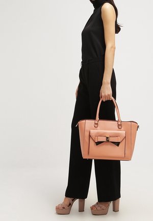 Borsa in ecopelle pesco con un accento di fiocco, forma strutturata, manici superiori e cerniere, abbinata a un outfit nero e scarpe con la zeppa.
