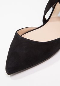 Scarpa piatta nera in suede con punta affusolata, foderata in beige e con una cinghia alla caviglia, su sfondo bianco.
