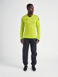 Limoengroene sportshirt met lange mouwen en zwarte accenten, gecombineerd met zwarte broek en zwarte sneakers. Bevat een logo op het shirt.